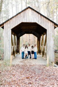 Five people and two dogs stand together on a wooden covered bridge in a wooded area with fallen leaves on the ground. The group is casually dressed and facing the camera. Trees without leaves are visible in the background.