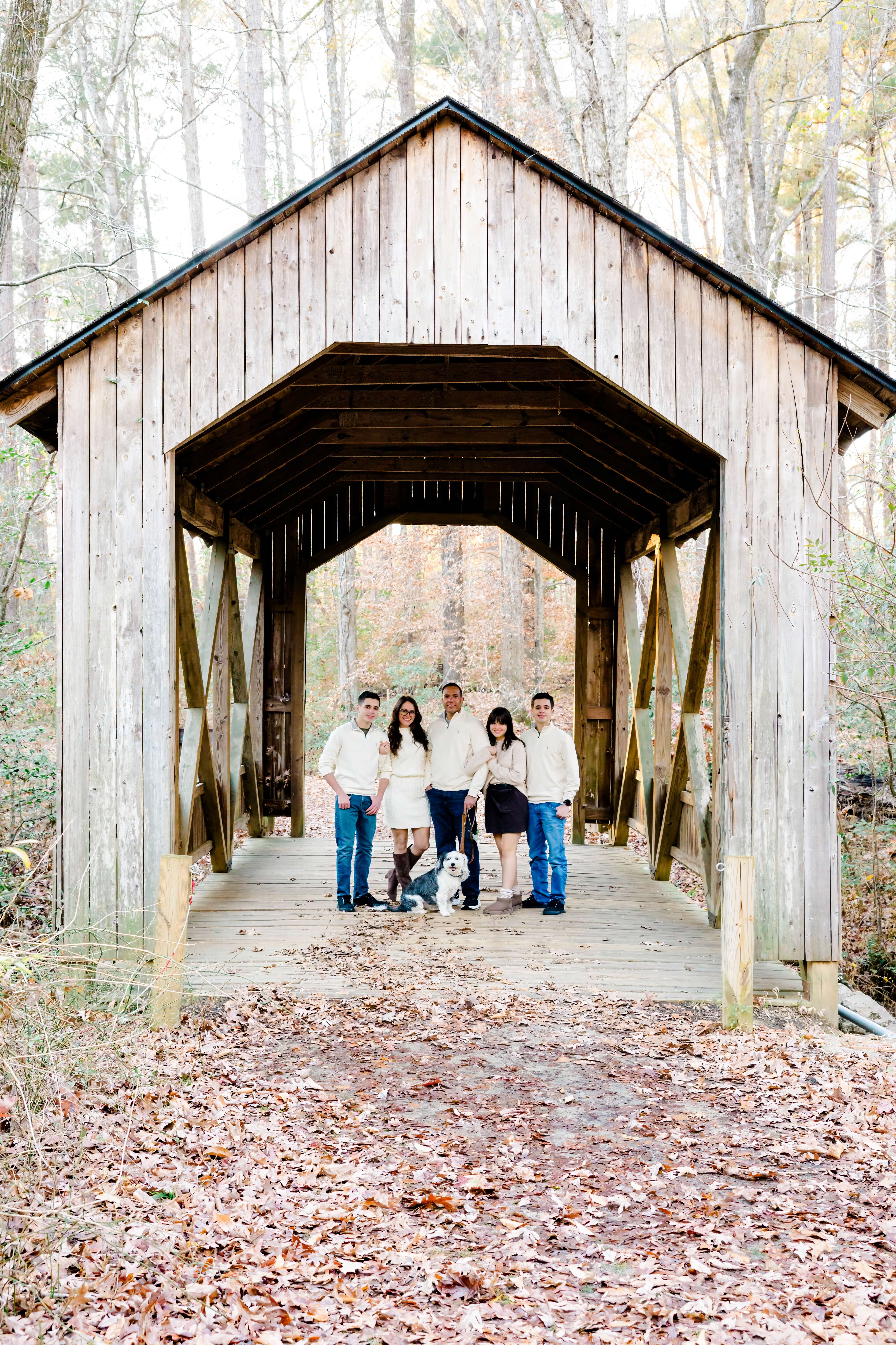 Five people and two dogs stand together on a wooden covered bridge in a wooded area with fallen leaves on the ground. The group is casually dressed and facing the camera. Trees without leaves are visible in the background.