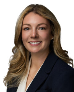 A woman with long blonde hair, wearing a dark blazer over a white blouse, smiles at the camera against a plain white background.