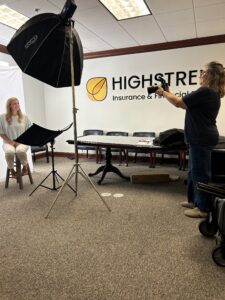 A photographer takes a picture of a woman sitting on a stool in an office with a "HIGHSTREET Insurance & Financial" sign on the wall. Studio lighting equipment is set up, and a table with items is in the background.