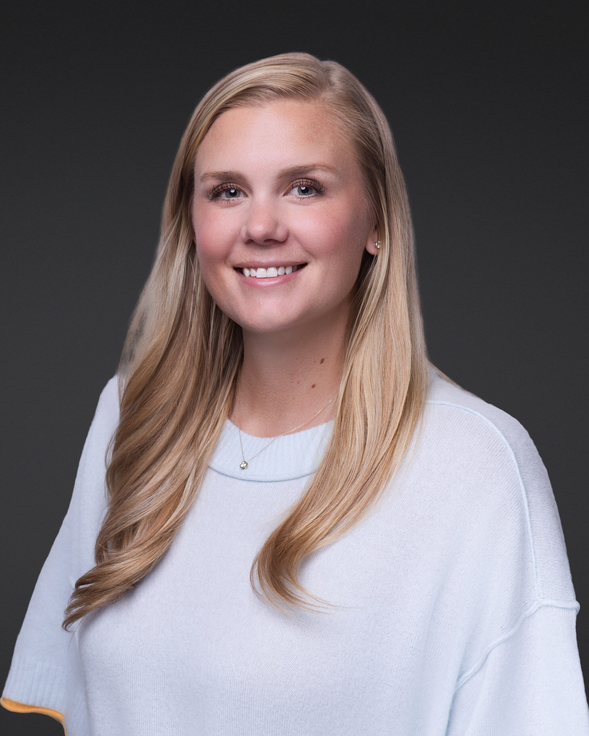 A woman with long blonde hair wearing a white sweater and a small pendant necklace is smiling and facing the camera against a plain dark background, showcasing the style of professional headshots in Fayetteville.