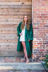 A young woman with long, wavy blonde hair wearing a green graduation gown over a short white dress stands against a brick wall and wooden door, smiling at the camera.