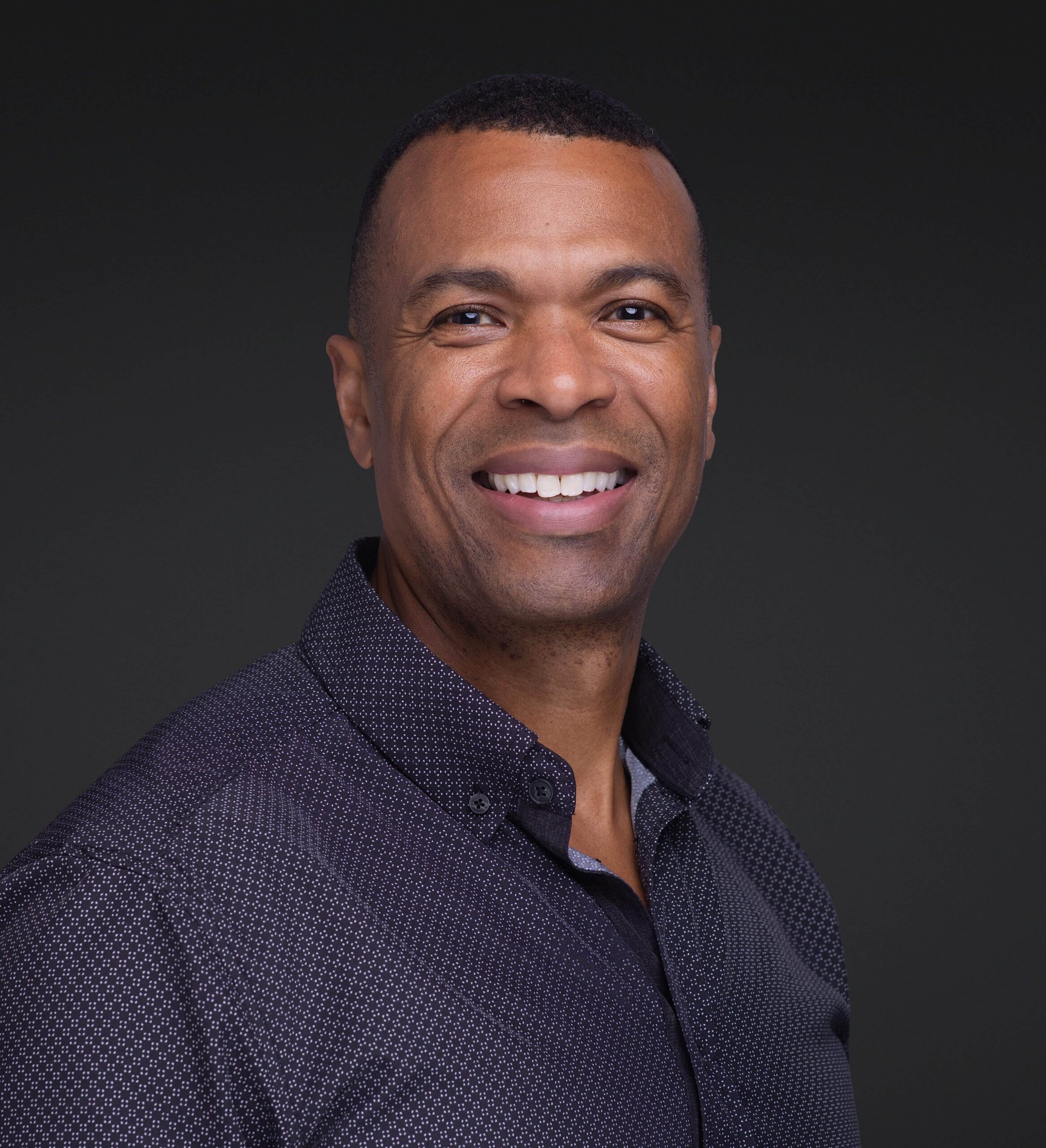 A man is smiling at the camera against a plain dark background. He is wearing a dark, patterned button-up shirt and has short hair—captured by a skilled headshot photographer in Southern Pines, NC.