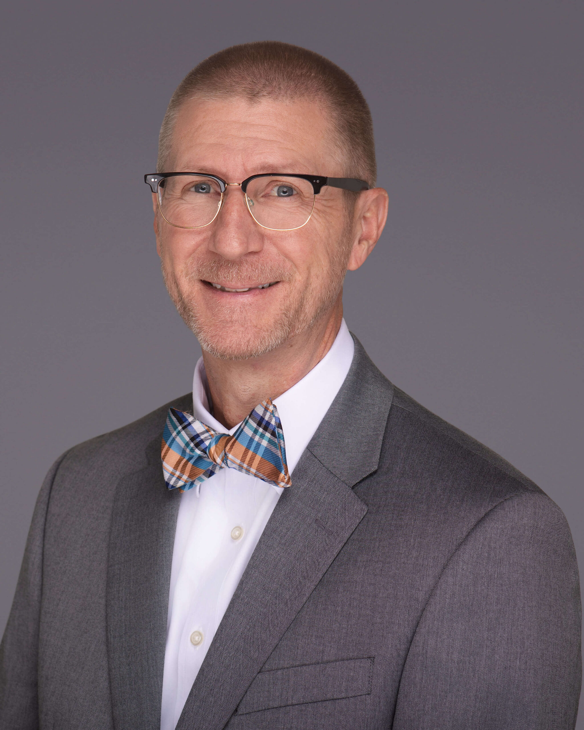 A man wearing glasses, a grey suit jacket, a white shirt, and a plaid bow tie smiles at the camera against a plain gray background—perfect for anyone seeking a professional headshot photographer in Southern Pines NC.