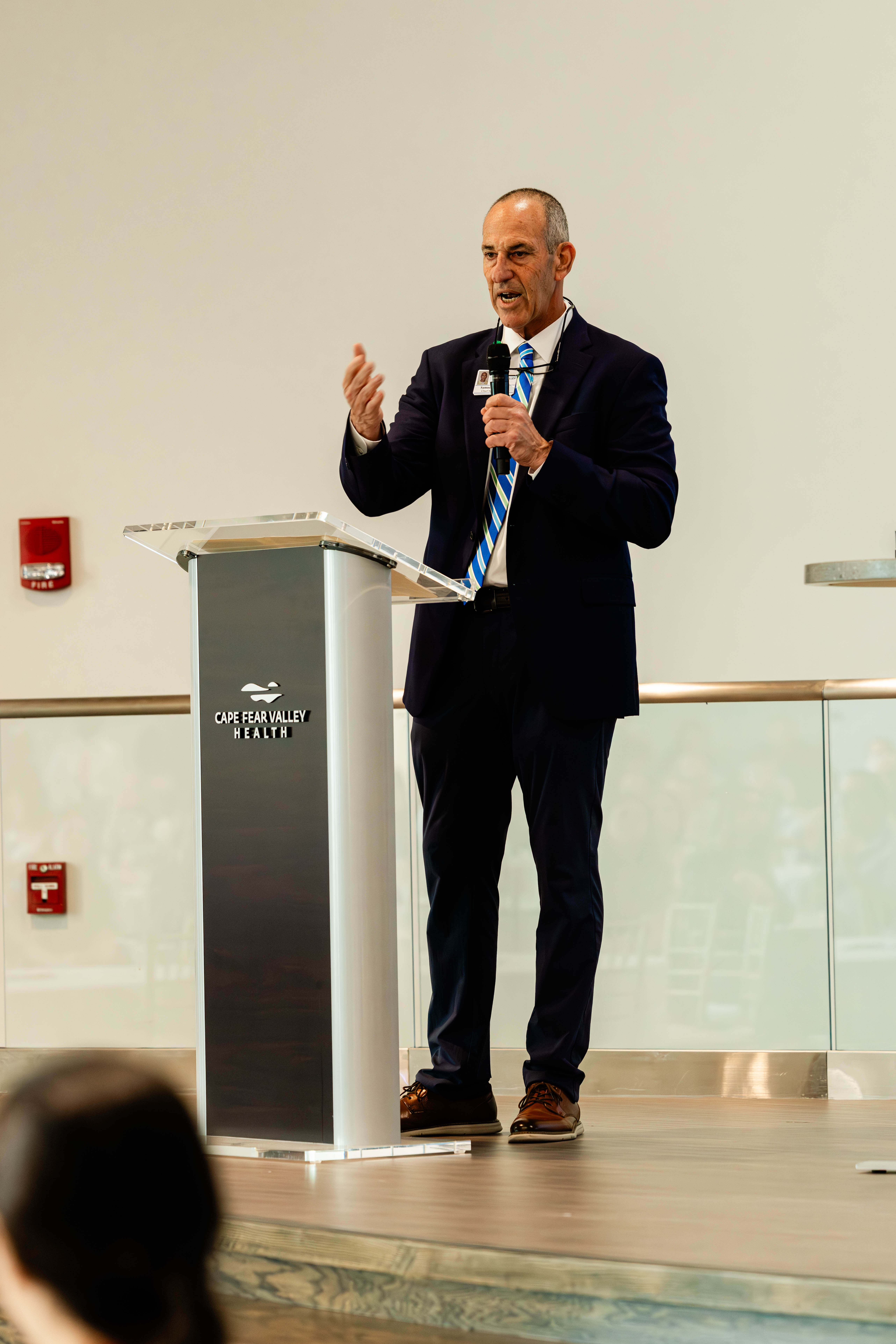 A man in a suit and striped tie speaks at a podium labeled “Cape Fear Valley Health,” holding a microphone in his left hand, with a white wall and glass railing in the background—an excellent example of corporate event photography Fayetteville NC.