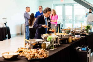A man in a suit serves himself food from a buffet table with various dishes, while several people stand and talk in the background. Captured in a bright, modern indoor event space, this image perfectly showcases the sophistication and energy of corporate event photography in Fayetteville NC.