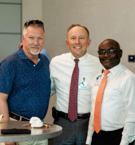 Three men stand side by side indoors, smiling at the camera during a corporate event. One wears a blue polo shirt, while the others are dressed in crisp white dress shirts with ties and name badges—ideal subjects for corporate event photography in Fayetteville, NC. A table with food and sunglasses is visible in the foreground.