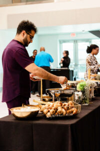 A man serves himself food from a buffet table with pastries and hot dishes during a corporate event. Other people stand and converse in the background of the modern indoor space, capturing the lively atmosphere typical of corporate event photography in Fayetteville, NC.