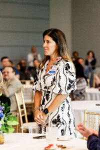A woman in a patterned dress stands by a round table at an indoor event, looking forward. Several people sit and observe in the background. Gold chairs and white tablecloths are visible, capturing the polished atmosphere of corporate event photography in Fayetteville, NC.