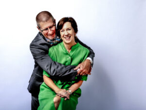 A man in a suit hugs a smiling woman in a green dress from behind as she sits on a stool, posed against a light background—an elegant moment captured during their personal branding photography in Fayetteville NC.
