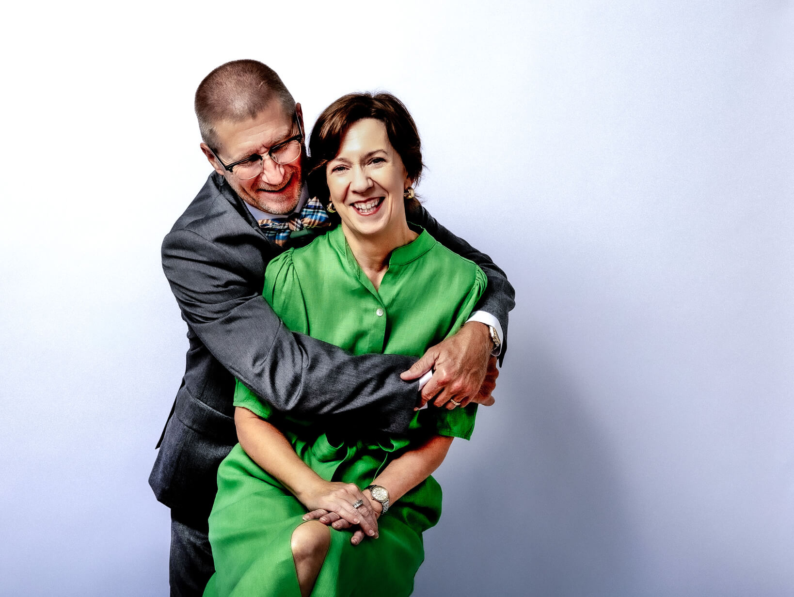 A man in a suit hugs a smiling woman in a green dress from behind as she sits on a stool, posed against a light background—an elegant moment captured during their personal branding photography in Fayetteville NC.