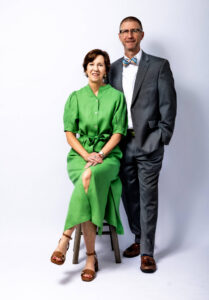 A woman in a green dress sits on a stool next to a man in a gray suit with a bow tie. Both are looking at the camera, standing against a plain white background—perfect for personal branding photography in Fayetteville NC.