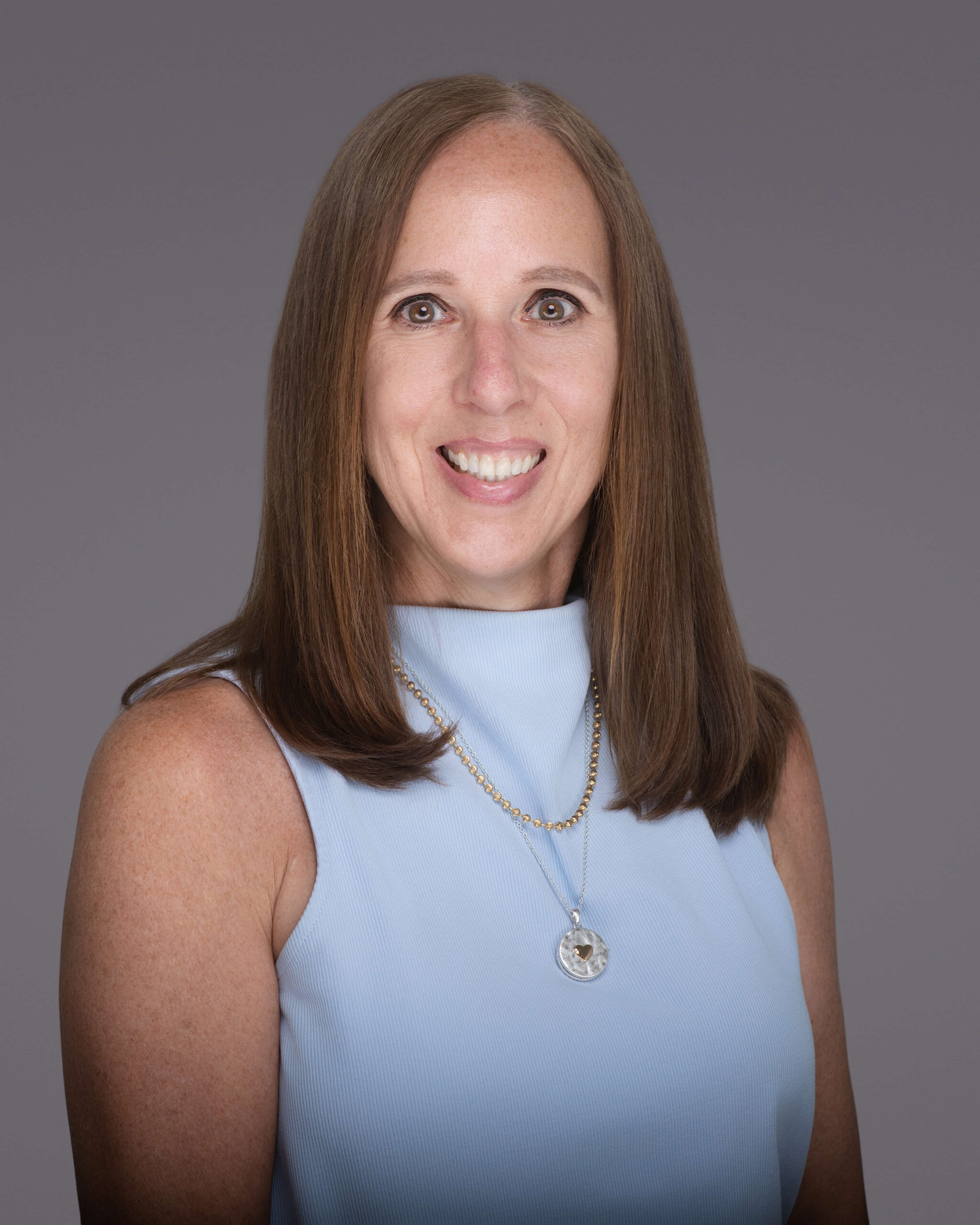 A woman with straight, shoulder-length brown hair wears a sleeveless light blue top and a gold necklace with a round pendant, smiling at the camera against a plain gray background—a polished portrait captured by a skilled headshot photographer in Sanford, NC.