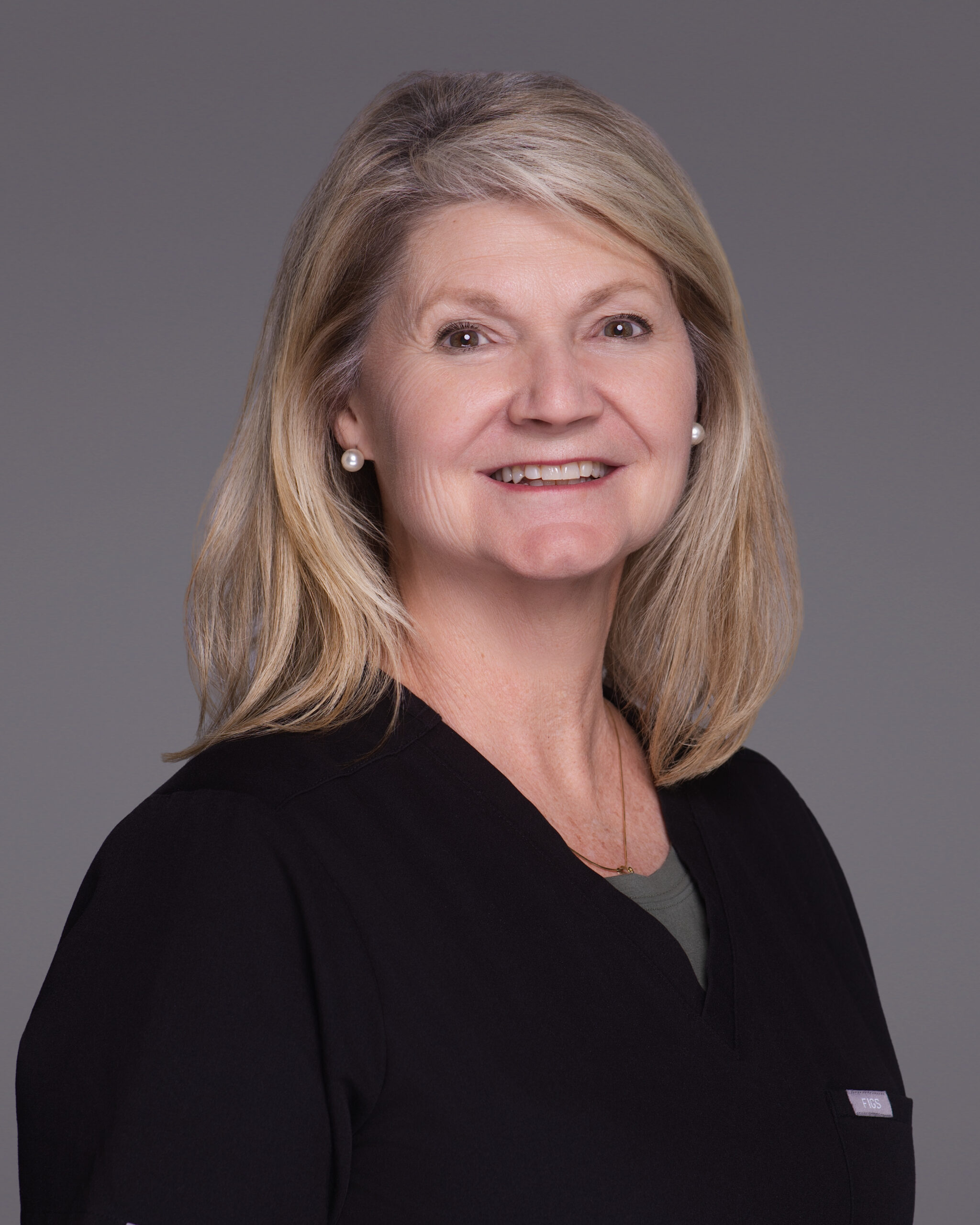 A middle-aged woman with shoulder-length blonde hair is smiling, wearing pearl earrings and a black top, posed against a plain gray background—an ideal example of professional healthcare headshots in Sandhills NC.
