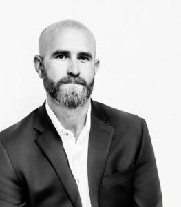A black and white portrait of a bald man with a beard wearing a suit jacket and white shirt.