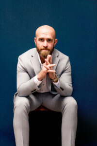 A bald man with a beard, wearing a grey suit, sits with his hands clasped against a blue background.