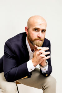 A bald man with a beard, wearing a white shirt and dark blazer, sits with his hands clasped under his chin.