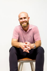 A smiling, bearded man wearing a pink polo shirt and dark trousers sits on a chair with his hands clasped.