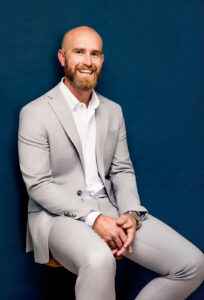 A smiling man with a beard, wearing a light gray suit and white button-down shirt, sitting against a dark blue background.